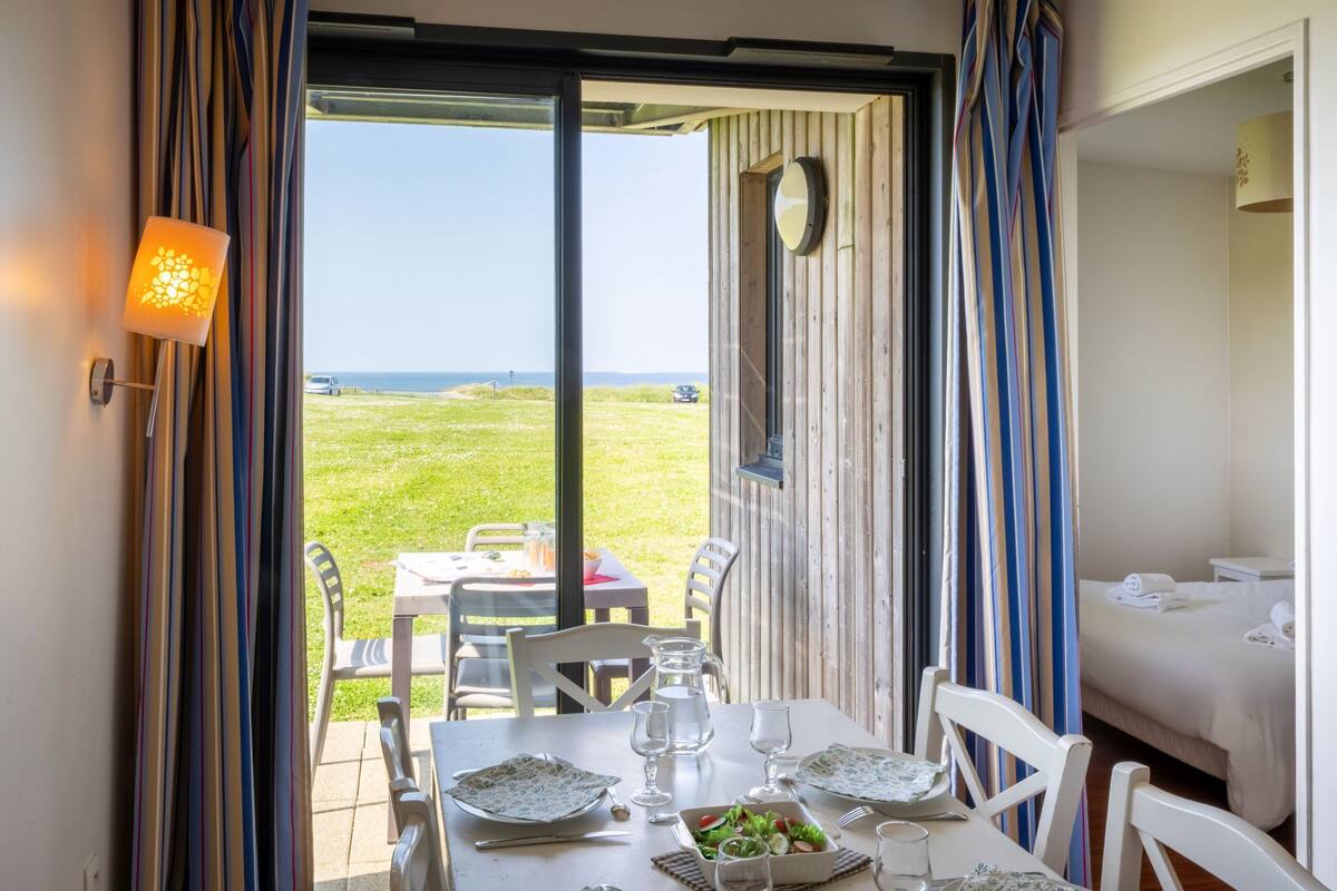A dining area with a table set for two, a view of the sea, and a bed in the adjacent room.