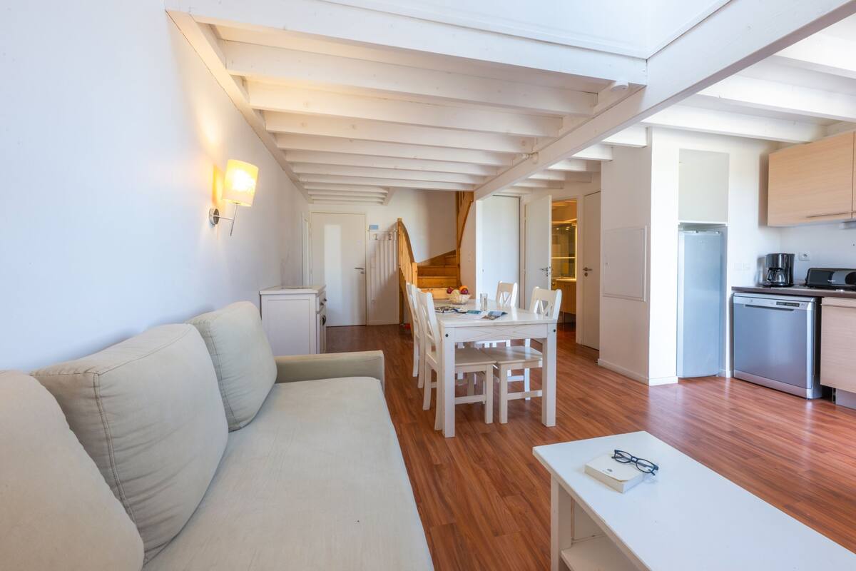 A modern, well-lit room with a wooden floor, a white sofa, a dining table with chairs, and a kitchen area with a refrigerator and coffee machine.