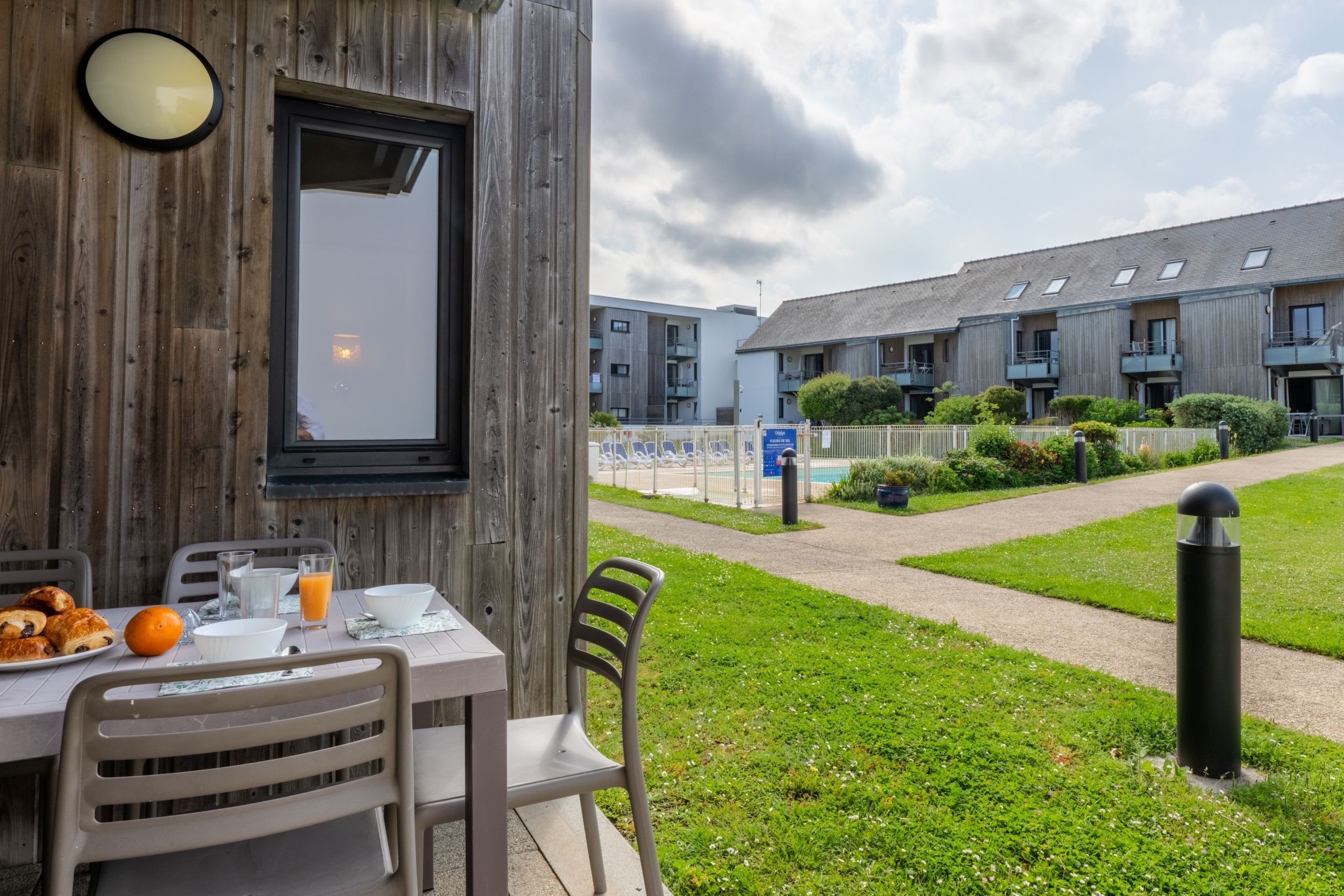 A breakfast table set with croissants, orange, and juice, outside a wooden building with a view of modern apartment buildings.