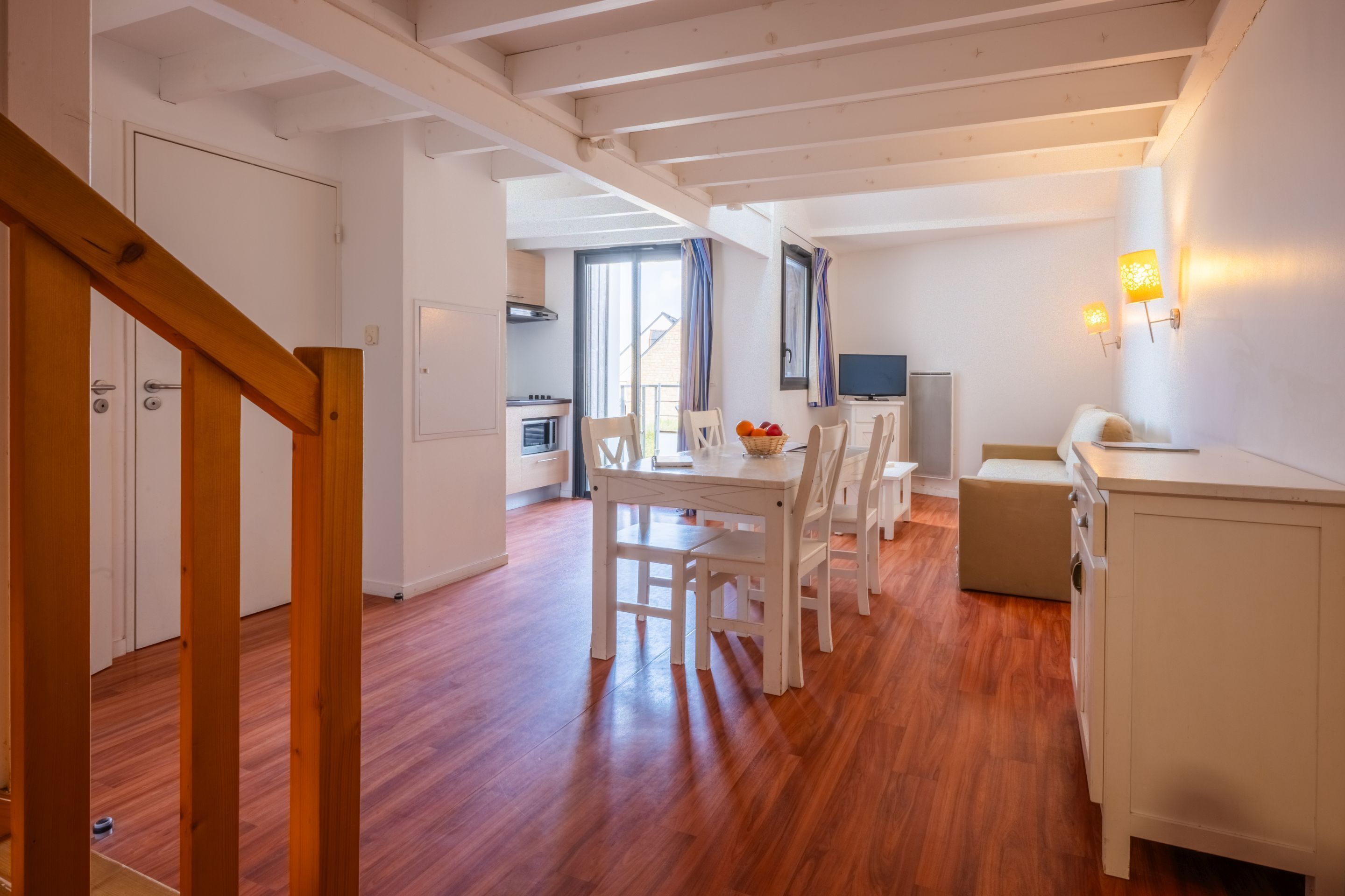A spacious living area with a wooden floor, white furniture, a dining table with chairs, a kitchen area, and a TV mounted on the wall.