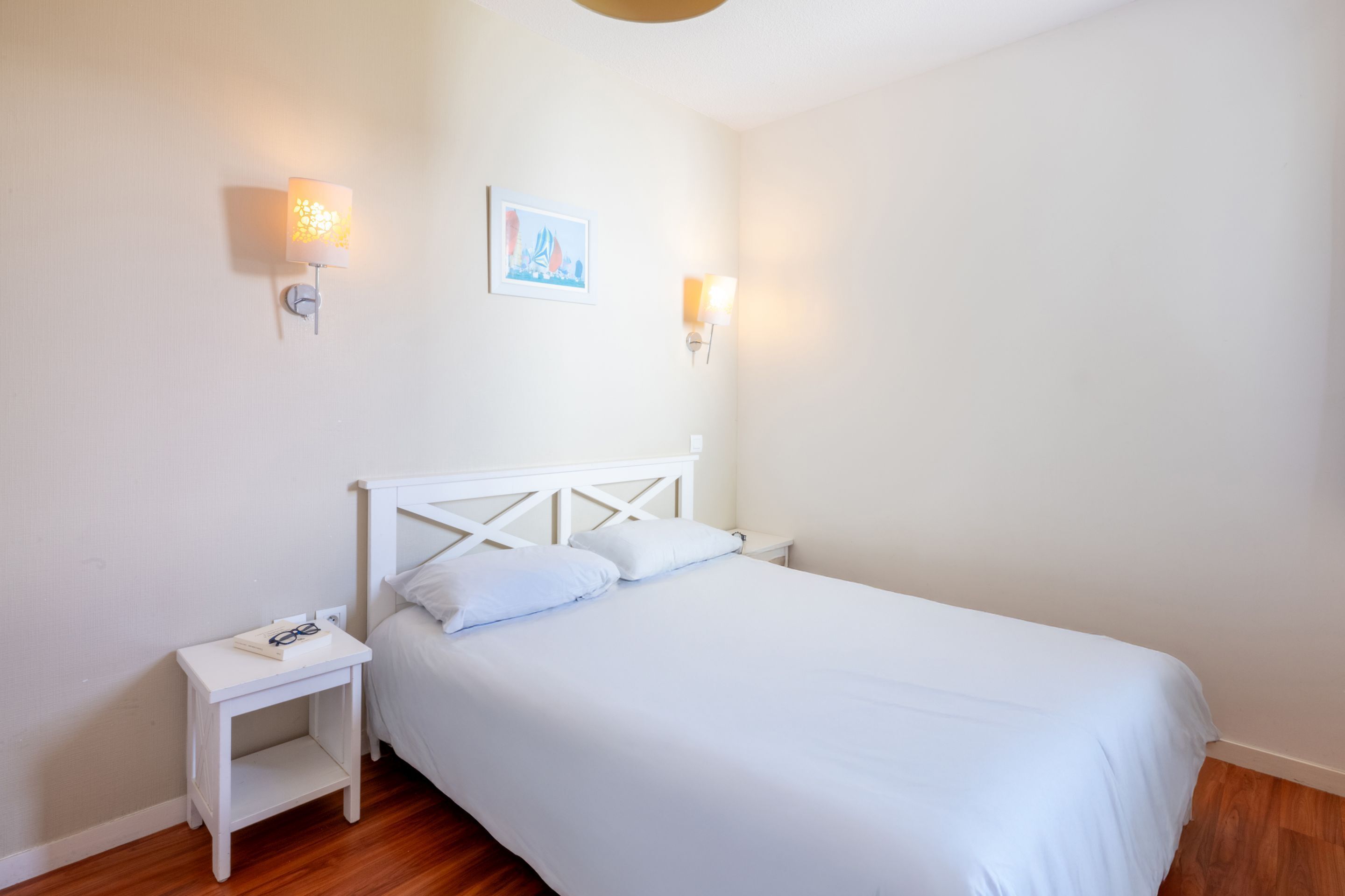 Un lit blanc avec une tête de lit en bois, une table de chevet avec une lampe et un tableau encadré au mur.