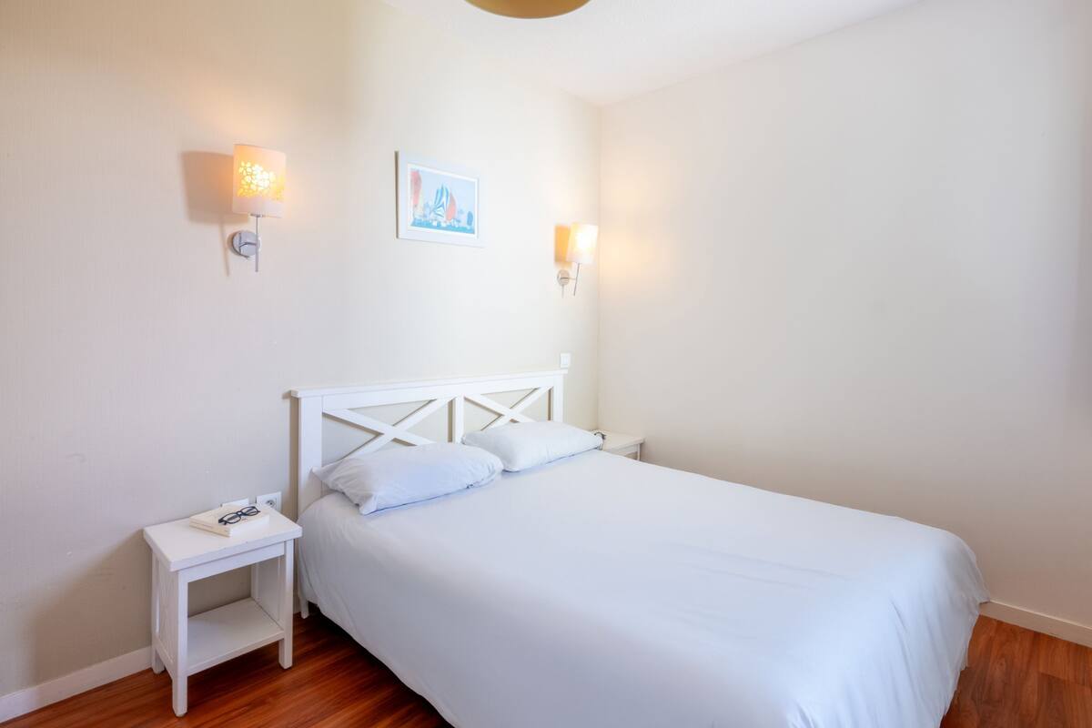 A bedroom with a white bed, two bedside tables, and wall-mounted lights.