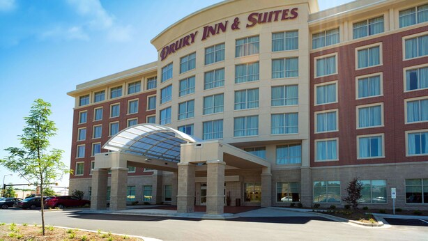 Un hotel Drury Inn & Suites con un diseño arquitectónico moderno, que destaca una entrada principal con toldo de cristal y varios pisos con amplias ventanas.