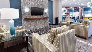 Un moderno vestíbulo de hotel con sofás a rayas, una mesa de centro de cristal, un televisor de pantalla plana montado en una pared con textura, y una zona de estar con vista al comedor.