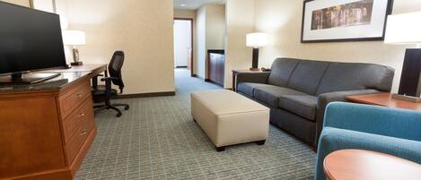 A hotel room with a flat-screen TV, a sofa, a loveseat, a wooden dresser, and a small table.