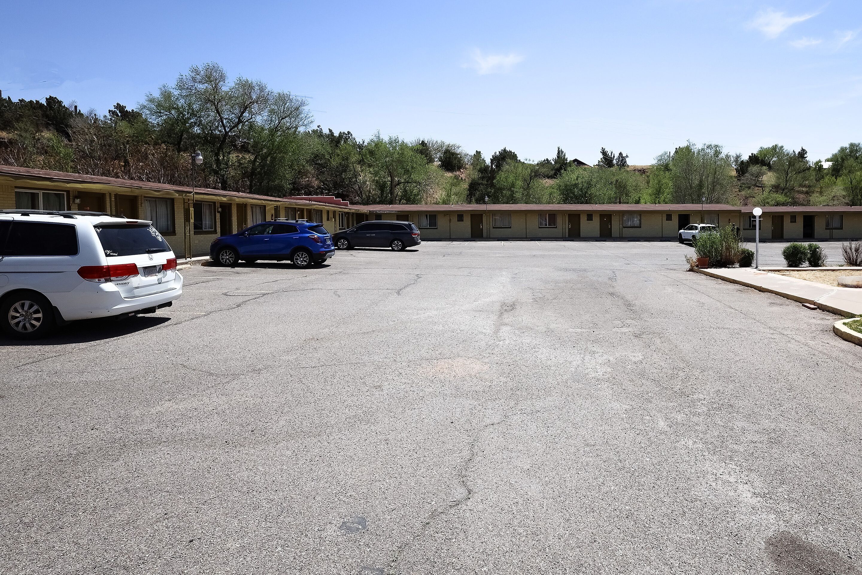 A parking lot with several cars, a motel with multiple rooms, and a hilly landscape with trees in the background.