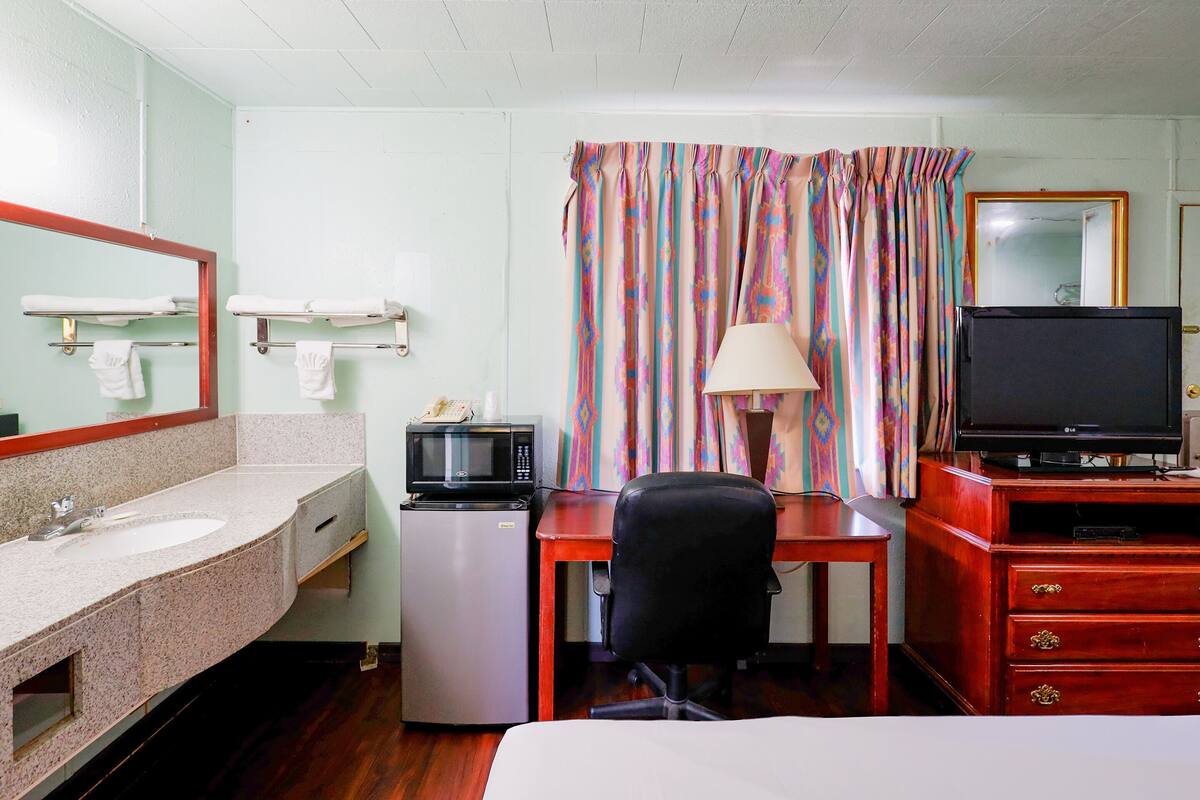 A hotel room with a bed, a desk with a chair, a television, a microwave, a refrigerator, a sink, and a mirror.
