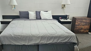 A bed with a grey quilt, two bedside tables with lamps, a wooden nightstand, and a wall clock.