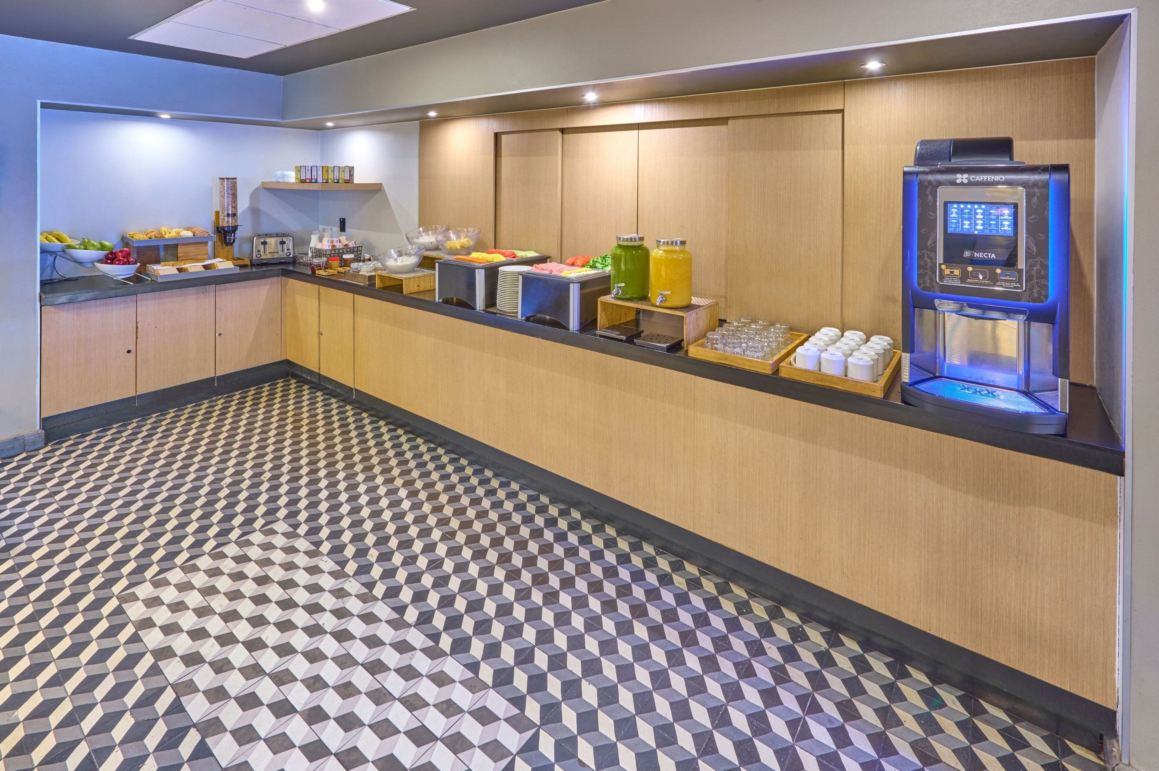 A hotel breakfast area with a self-service beverage station, a fruit bowl, and a variety of breakfast items on the counter.
