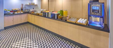 A hotel breakfast area with a self-service beverage station, a fruit bowl, and a variety of breakfast items on the counter.