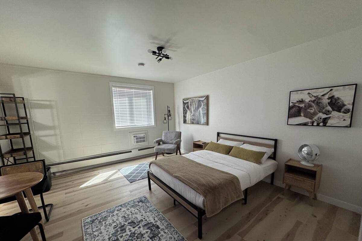 A bedroom with a bed, a chair, a desk, a standing fan, and artwork on the walls.