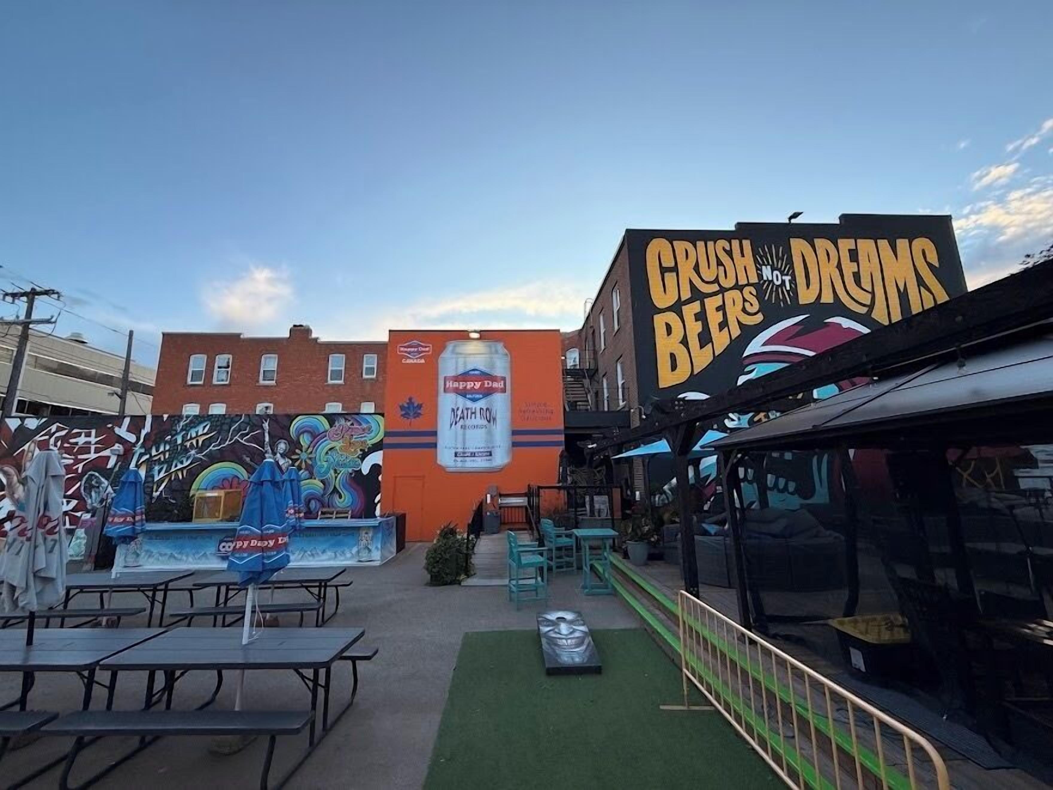 Un espace extérieur aménagé avec des tables et des chaises, une fresque sur le bâtiment et une enseigne indiquant « CRUSH BEERS & DREAMS ».