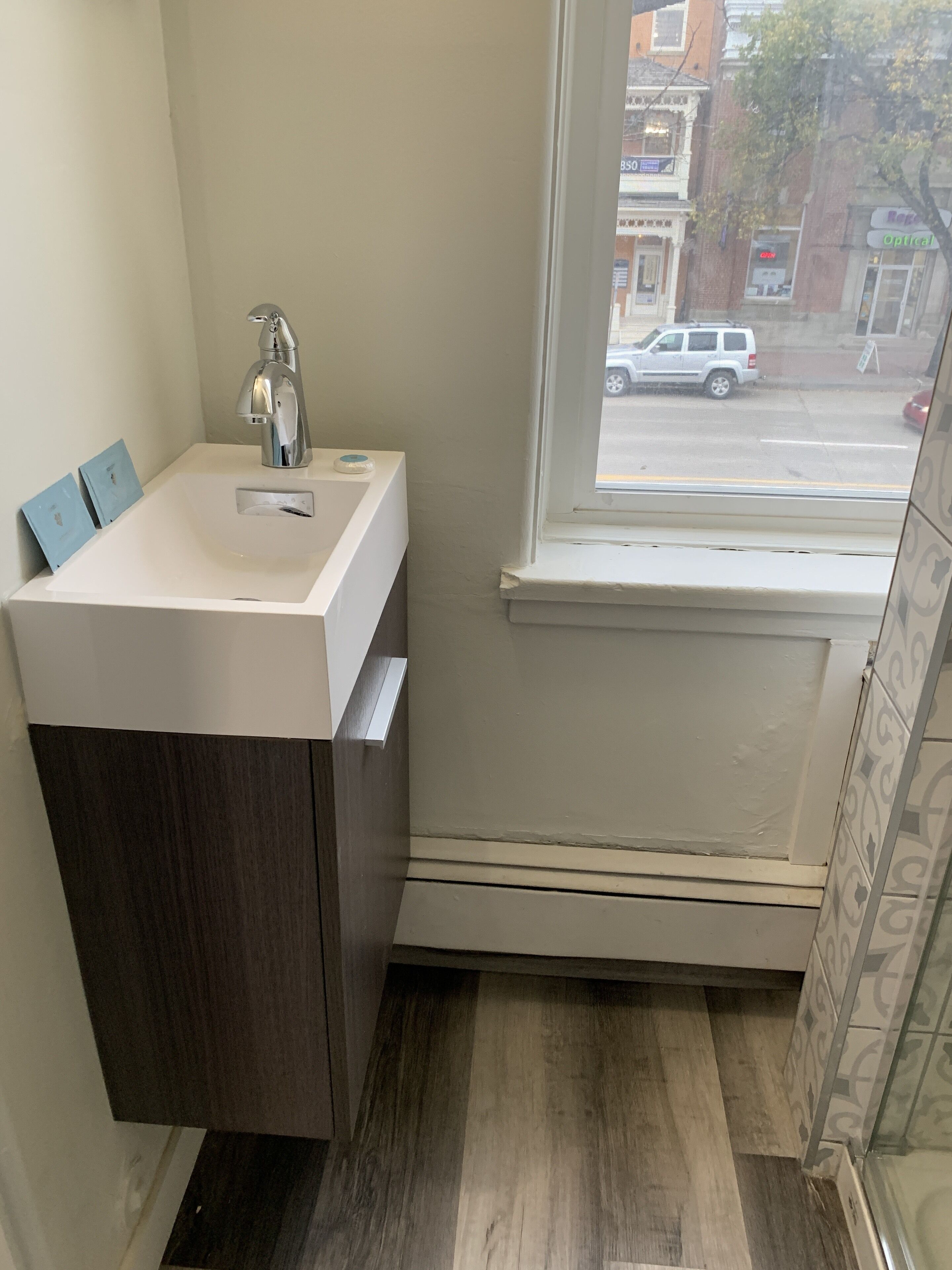 Une salle de bain avec un lavabo blanc, un meuble en bois foncé et une fenêtre donnant sur la rue.