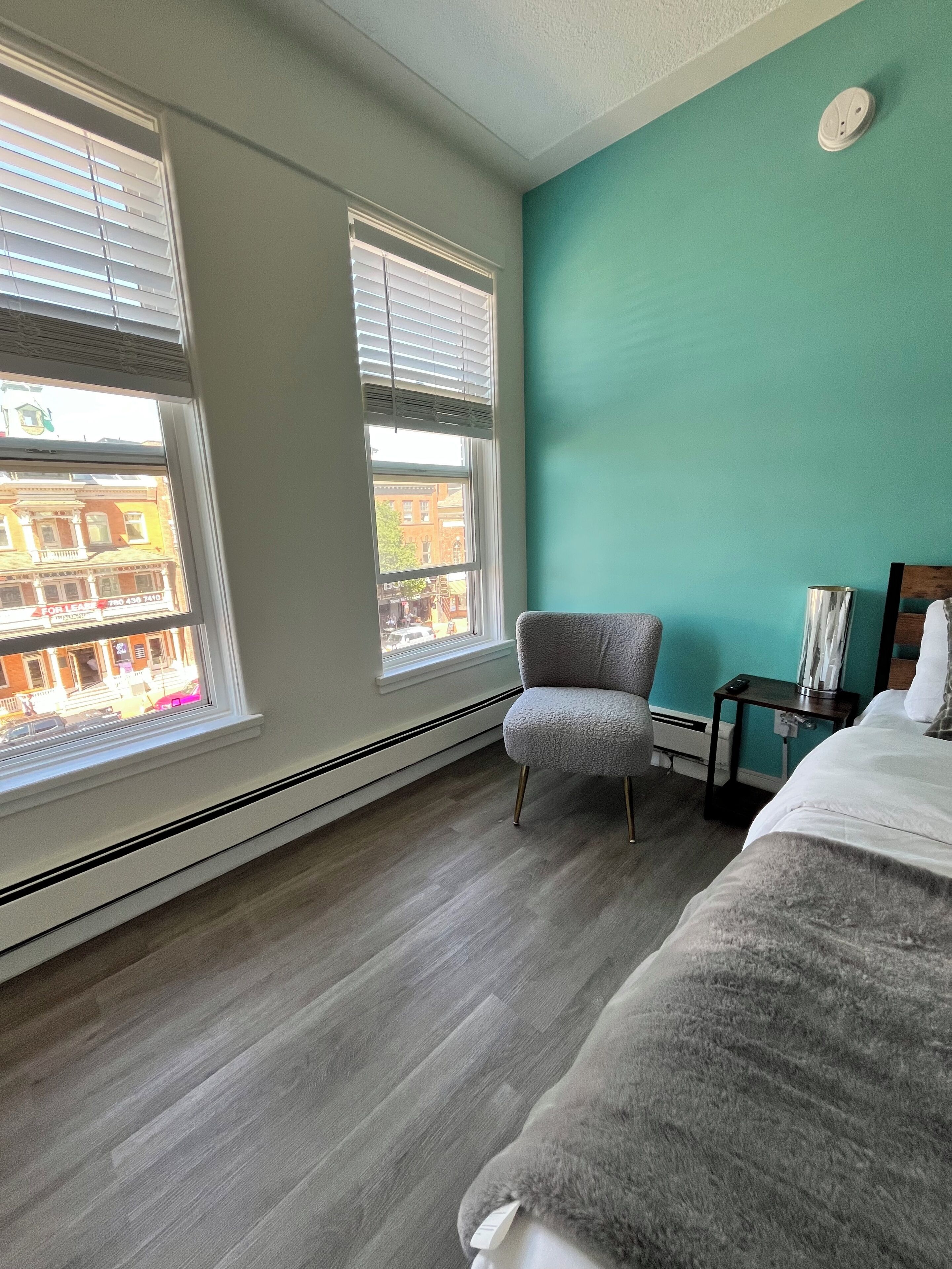 Una habitación de hotel con un ventanal grande, una silla gris, una cama y una mesita de noche.