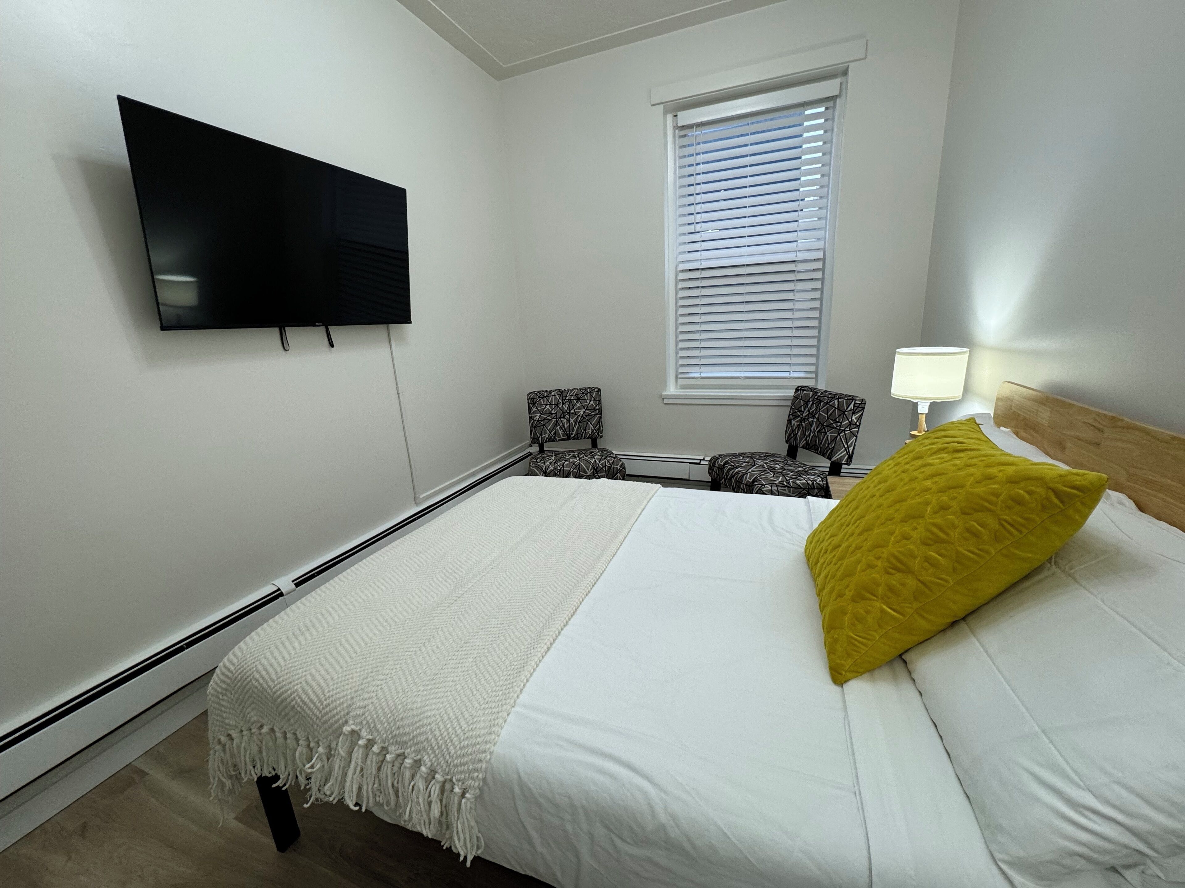 A hotel room with a bed, a TV mounted on the wall, two patterned chairs, a window with blinds, and a bedside lamp.