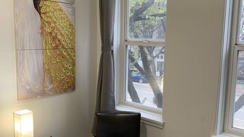 A room with a bed, a chair, a nightstand with a lamp, a window with a view of trees and a car, and a peacock painting on the wall.