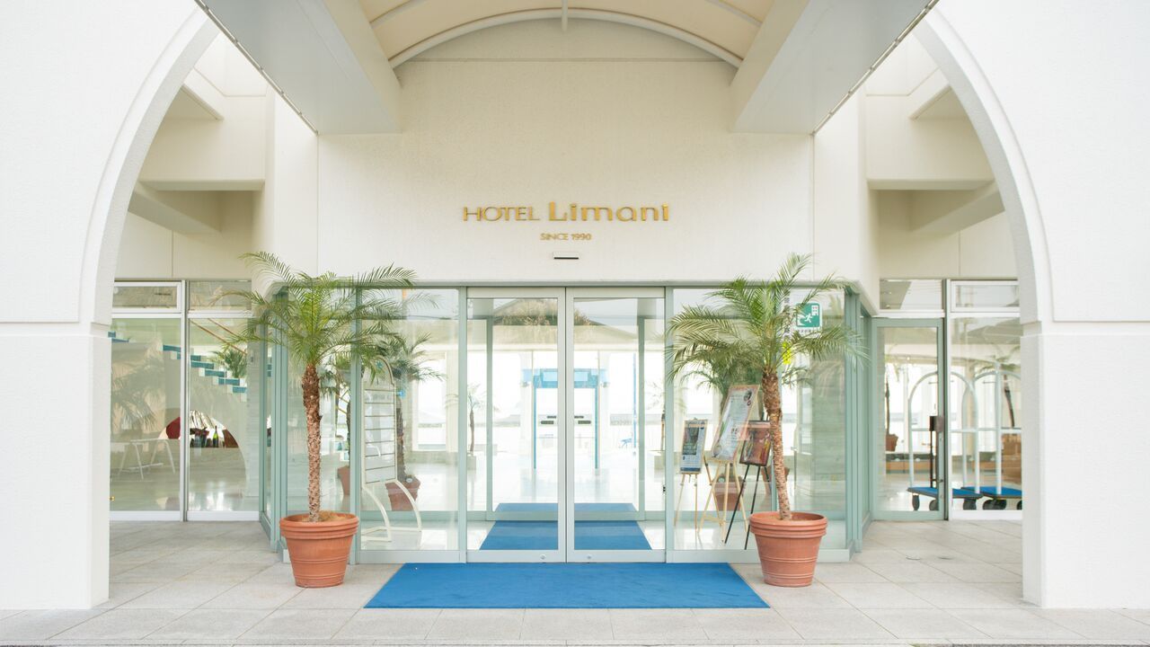 La entrada del Hotel Limani, con fachada de vidrio, palmeras y una alfombra azul que conduce a la entrada.
