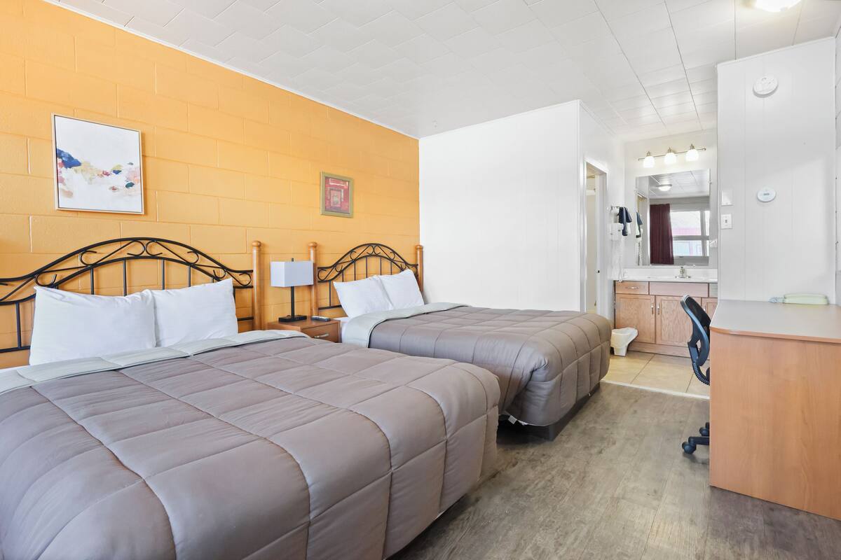 Une chambre d’hôtel avec deux lits, un bureau, un miroir et une salle de bain visible à travers une porte ouverte.