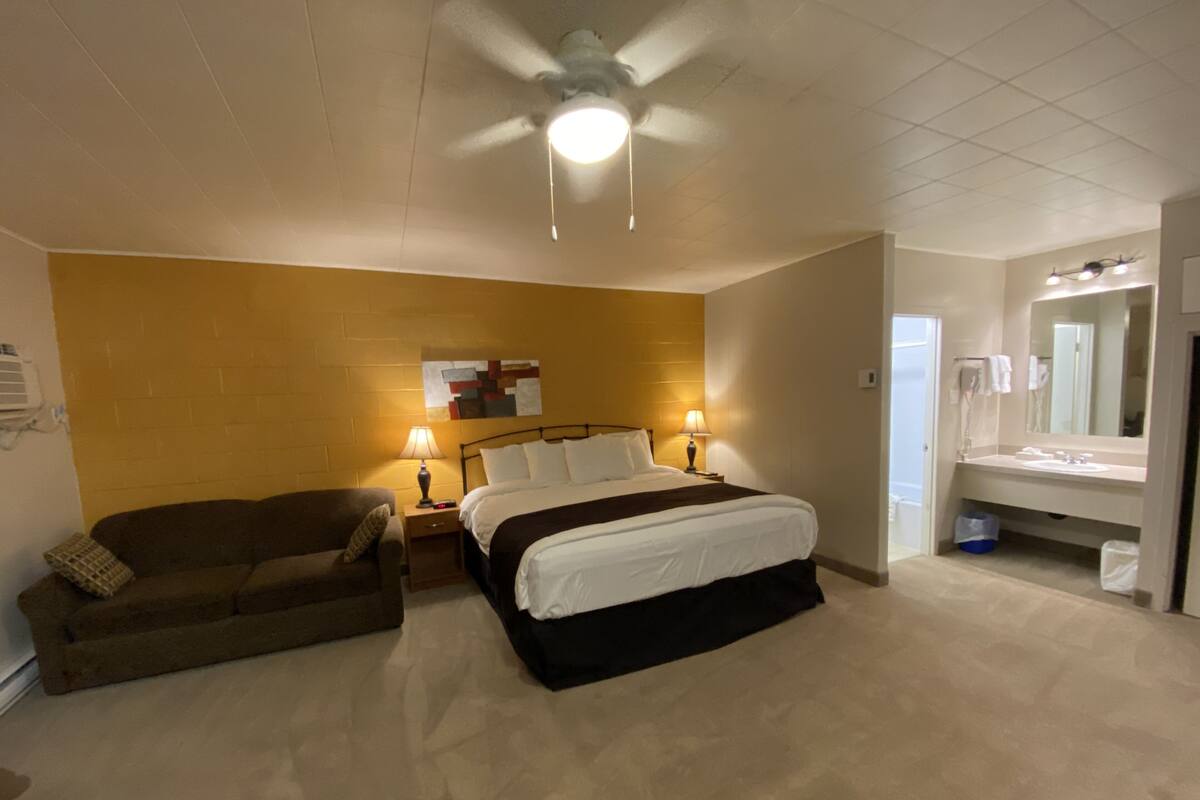 Une chambre d’hôtel comprenant un lit, une table de chevet, une lampe, un ventilateur de plafond, une salle de bain avec un lavabo et un miroir, et un climatiseur fixé au mur.