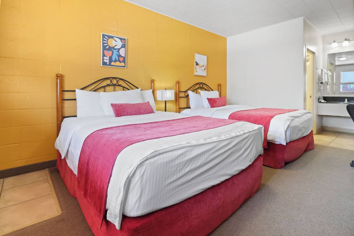 Une chambre d’hôtel avec deux lits, chacun doté de draps blancs et rouges, d’une tête de lit en bois et d’un mur jaune. On y trouve un bureau avec une chaise et un miroir.