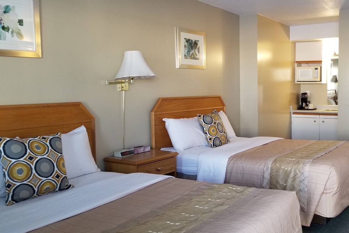 A hotel room with two beds, a bedside table with a lamp, a small desk, and a kitchenette area.