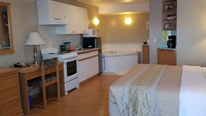 A compact bedroom with a bed, bedside table, and a small desk with a lamp. Adjacent is a kitchenette with a stove, microwave, and sink. - Athabasca Lodge Motel (Athabasca)