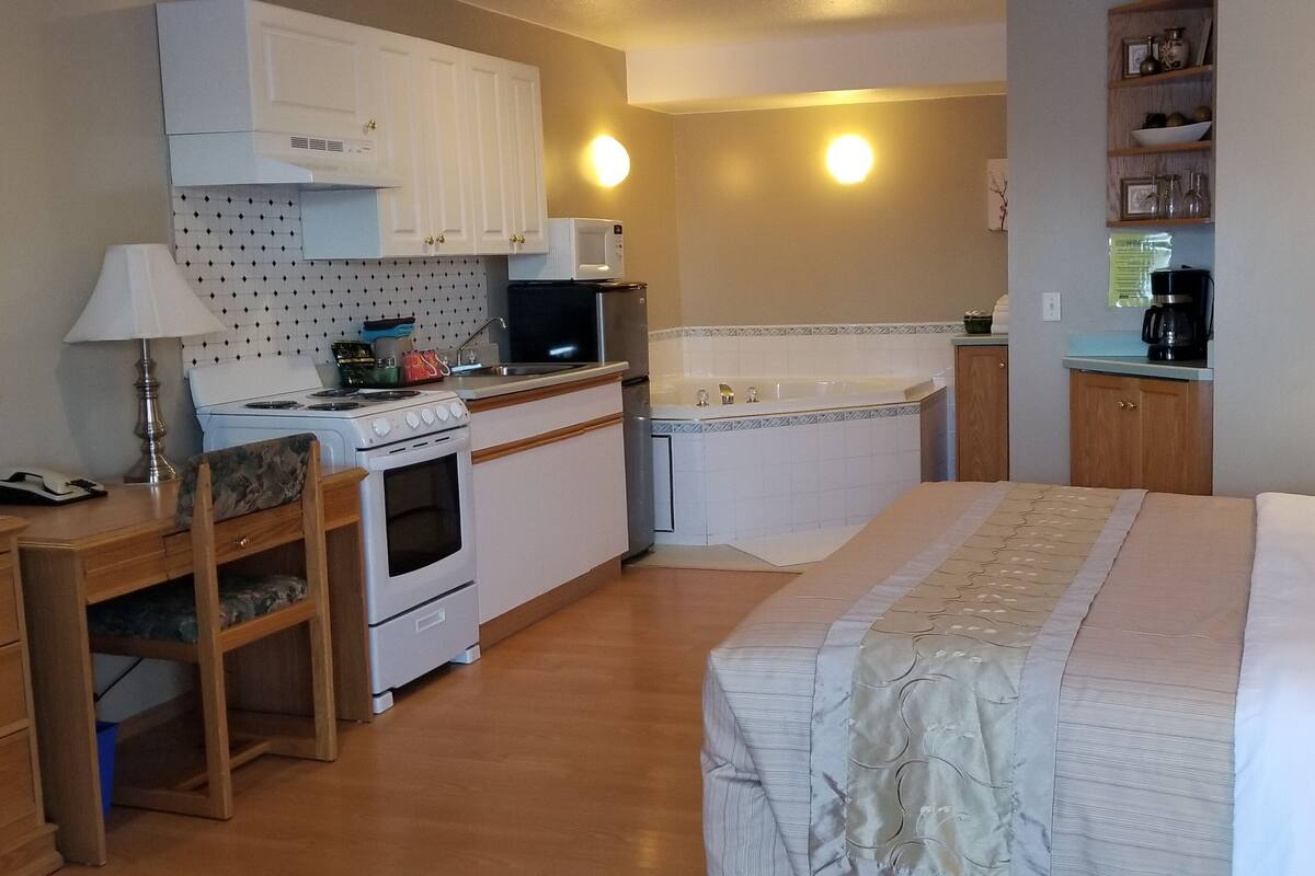 A compact bedroom with a bed, bedside table, and a small desk with a lamp. Adjacent is a kitchenette with a stove, microwave, and sink.