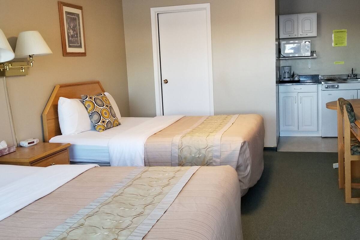 A hotel room with two beds, a desk, a chair, a nightstand, a lamp, a microwave, and a small kitchenette.