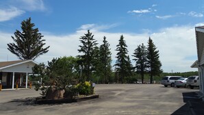 A parking lot with several vehicles, surrounded by tall evergreen trees and a building with a sloped roof. - Athabasca Lodge Motel (Athabasca)