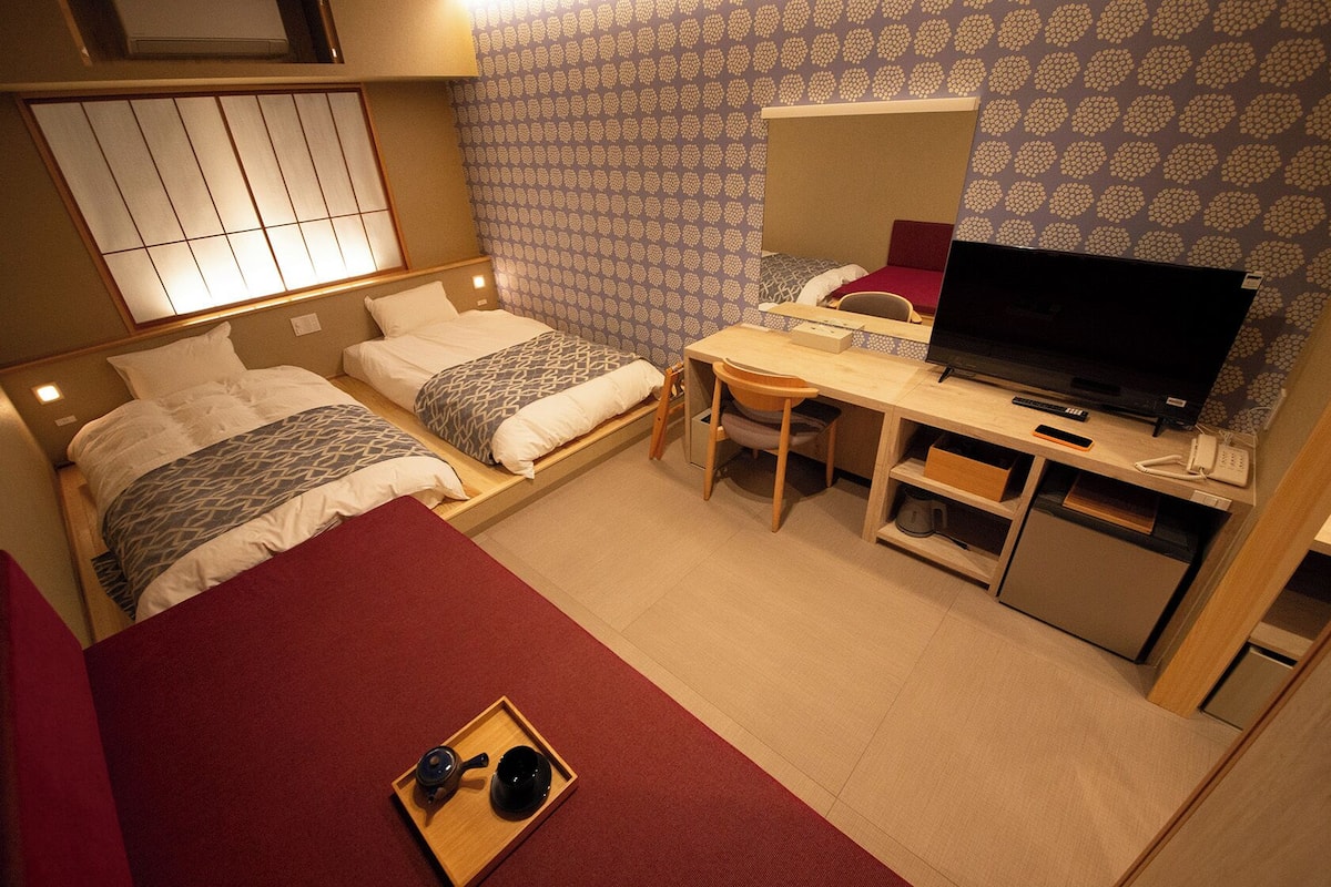 A hotel room with two beds, a desk with a TV, a small kitchenette, and a sliding shoji screen.
