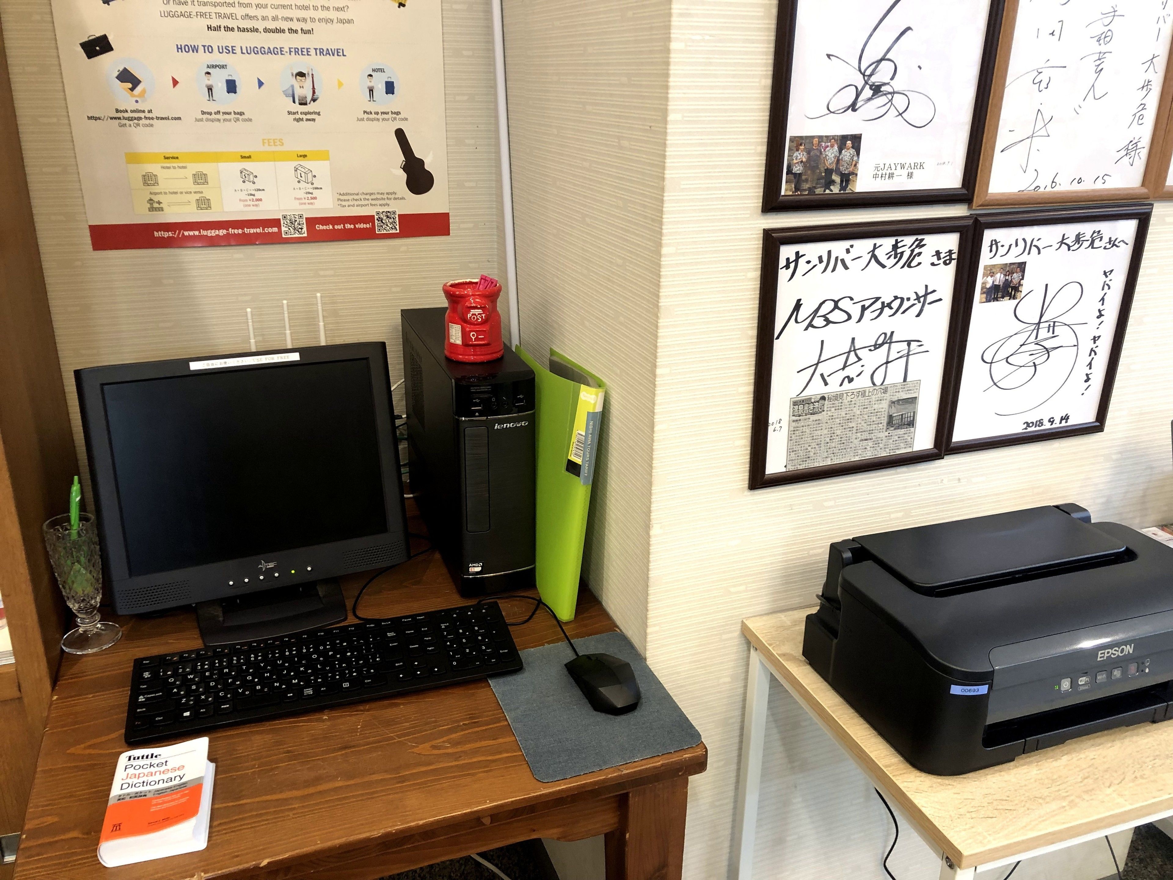 A desk with a computer, keyboard, mouse, and a printer. There are framed pictures on the wall and a small red object on the desk.