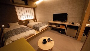 Habitación de hotel compacta con dos camas, televisión, pequeña kitchenette y una mesa redonda con tetera y taza.