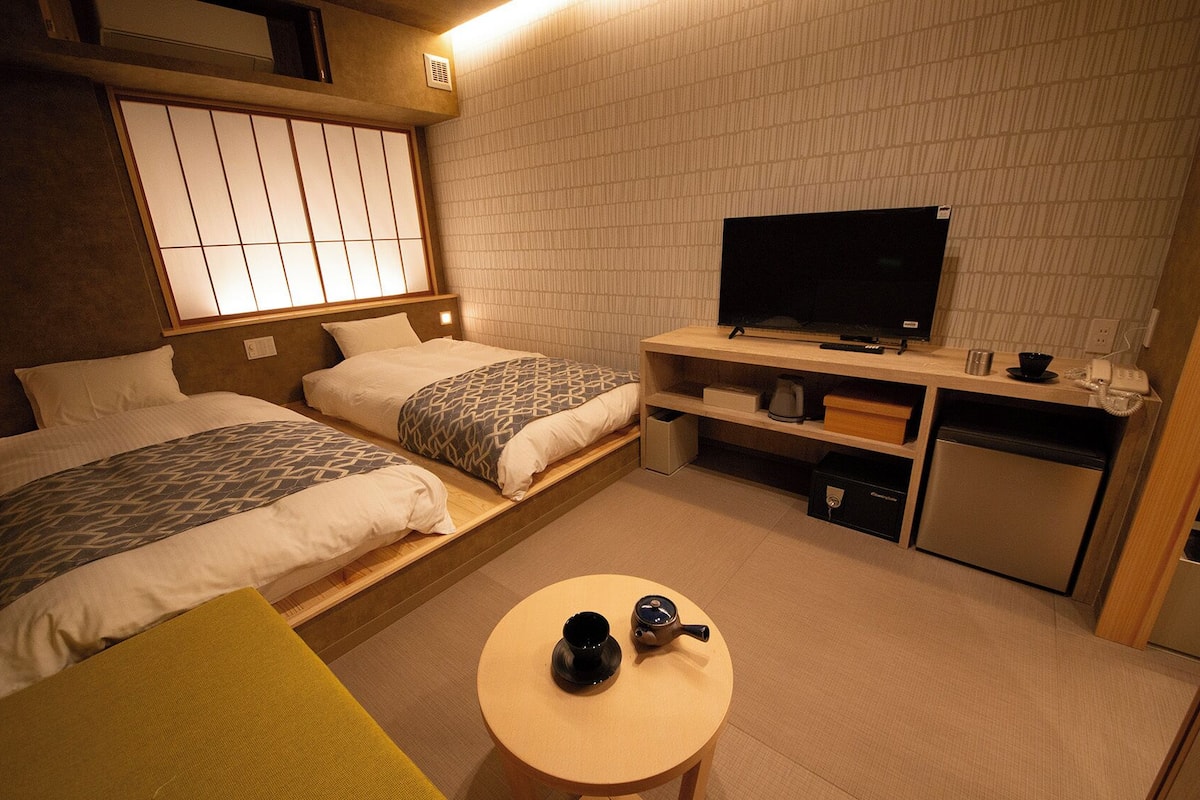 A compact hotel room with two beds, a television, a small kitchenette, and a round table with a teapot and cup.