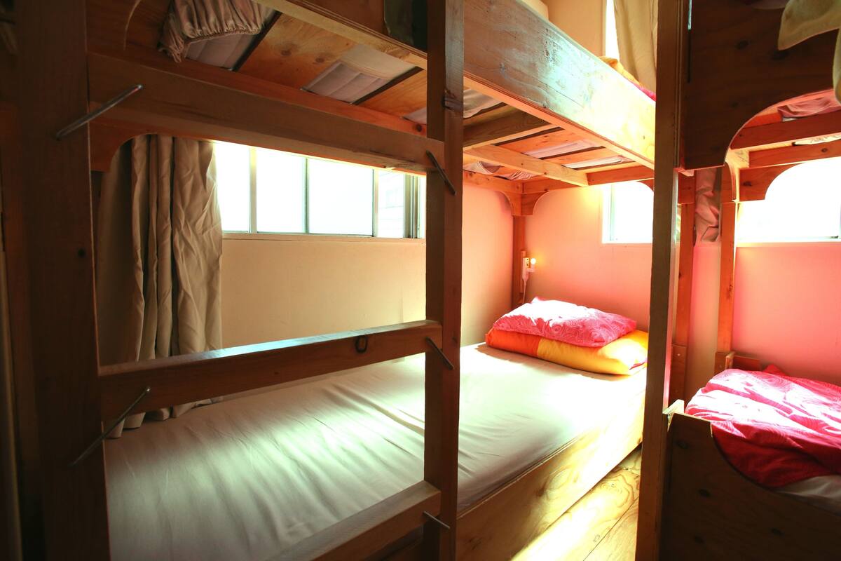 A bunk bed room with wooden construction, a window with curtains, and a bed with a pink and orange pillow.
