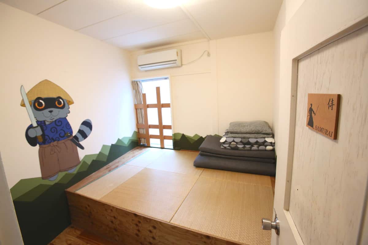 A room with tatami flooring, a futon bed, a wall-mounted air conditioner, and a door with a 'SAMURAI' sign.