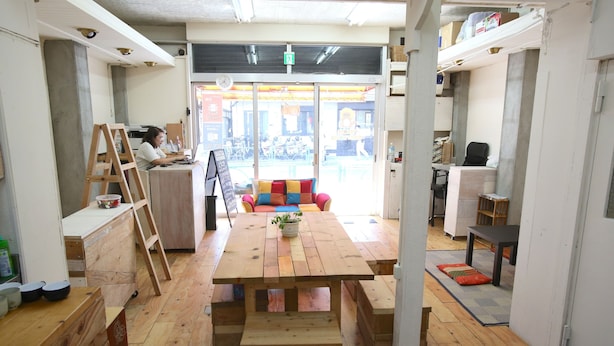 A wooden interior with a central table, benches, and a person working at a desk. - Yadoya Guesthouse Green - Hostel (Tokyo)