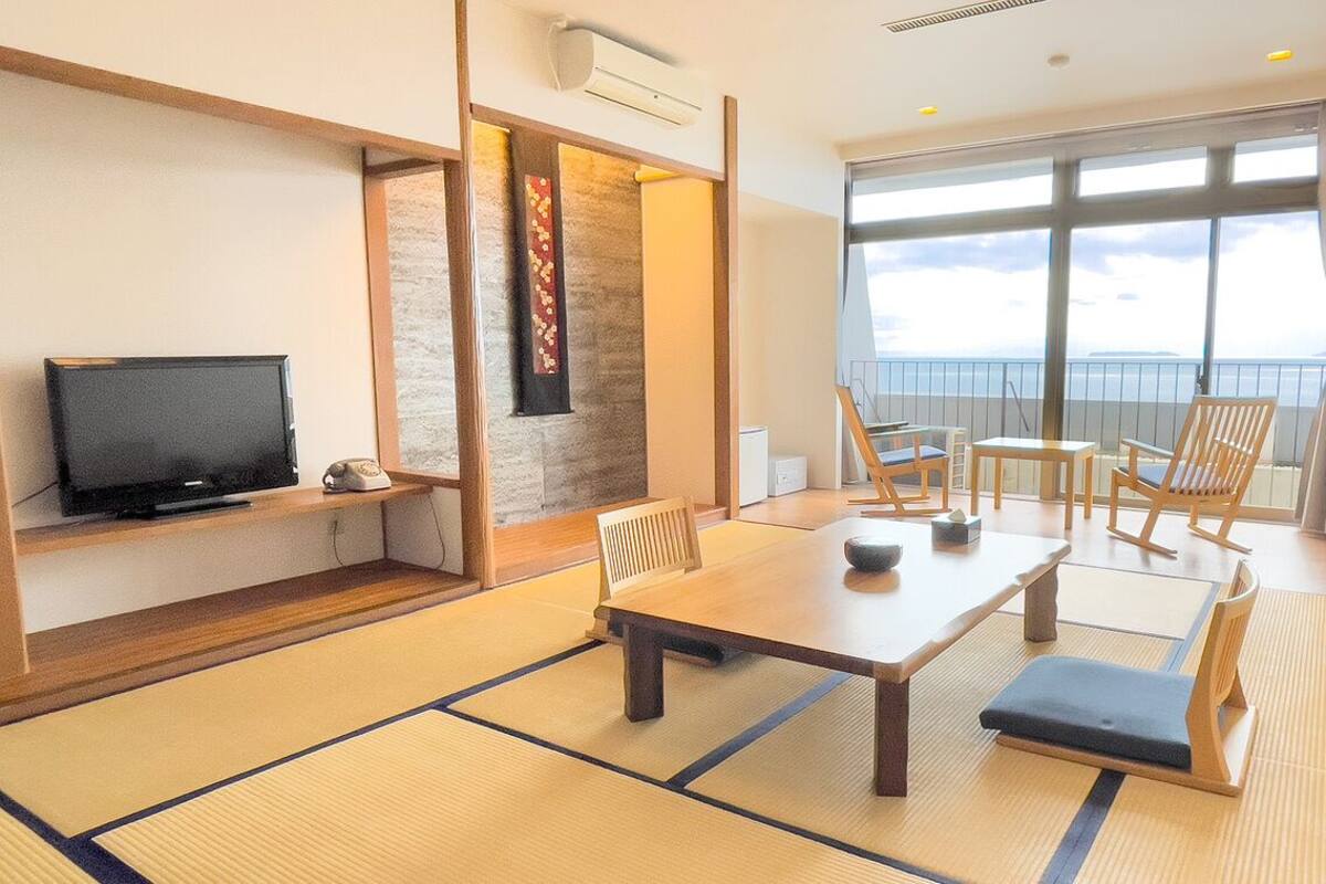 Ein Zimmer mit Tatami-Matten, einem Fernseher, einem Esstisch, Stühlen und Meerblick.