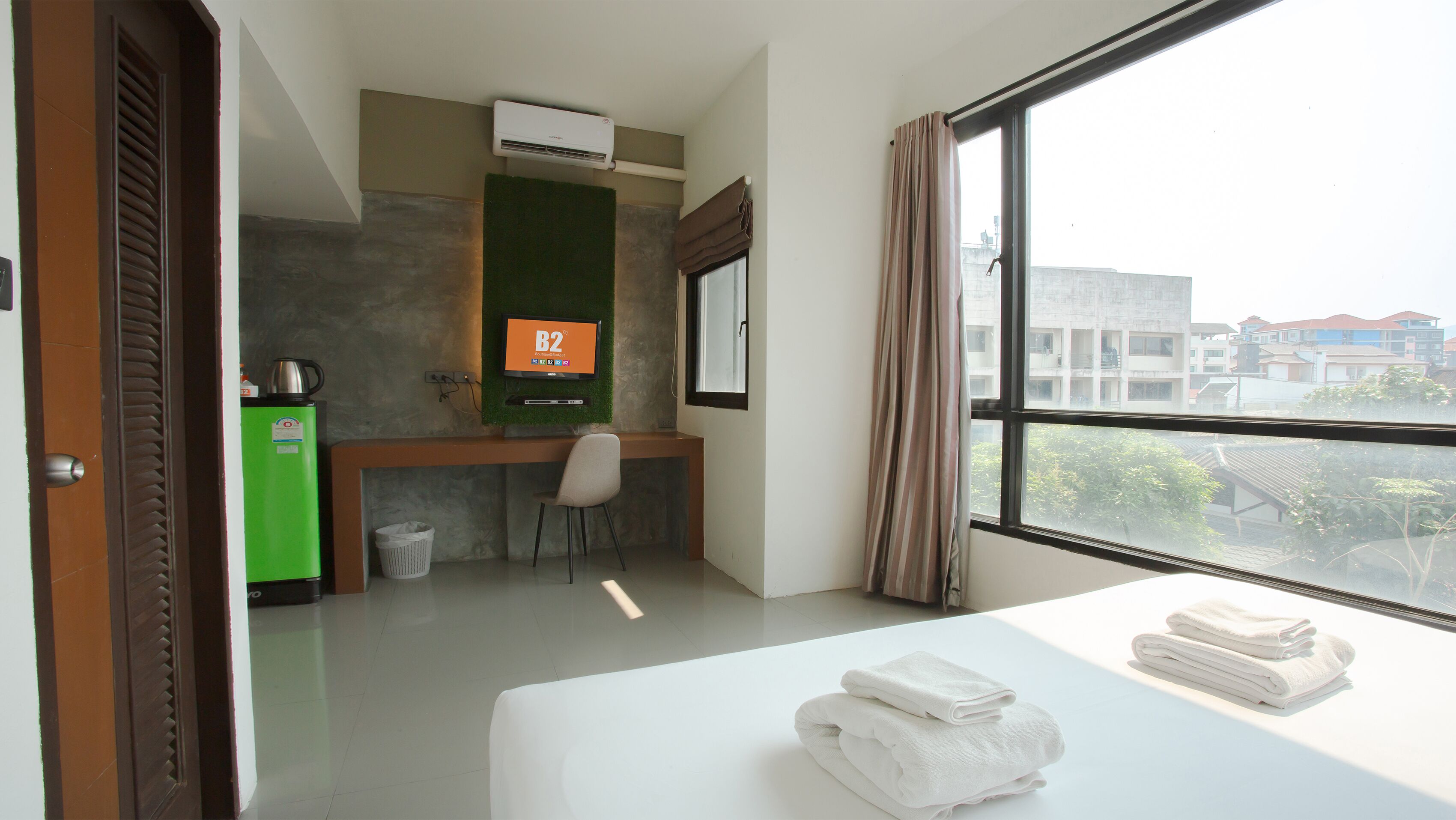 A hotel room with a bed, a desk, a chair, a green minibar, a trash bin, and a window with a view of buildings.