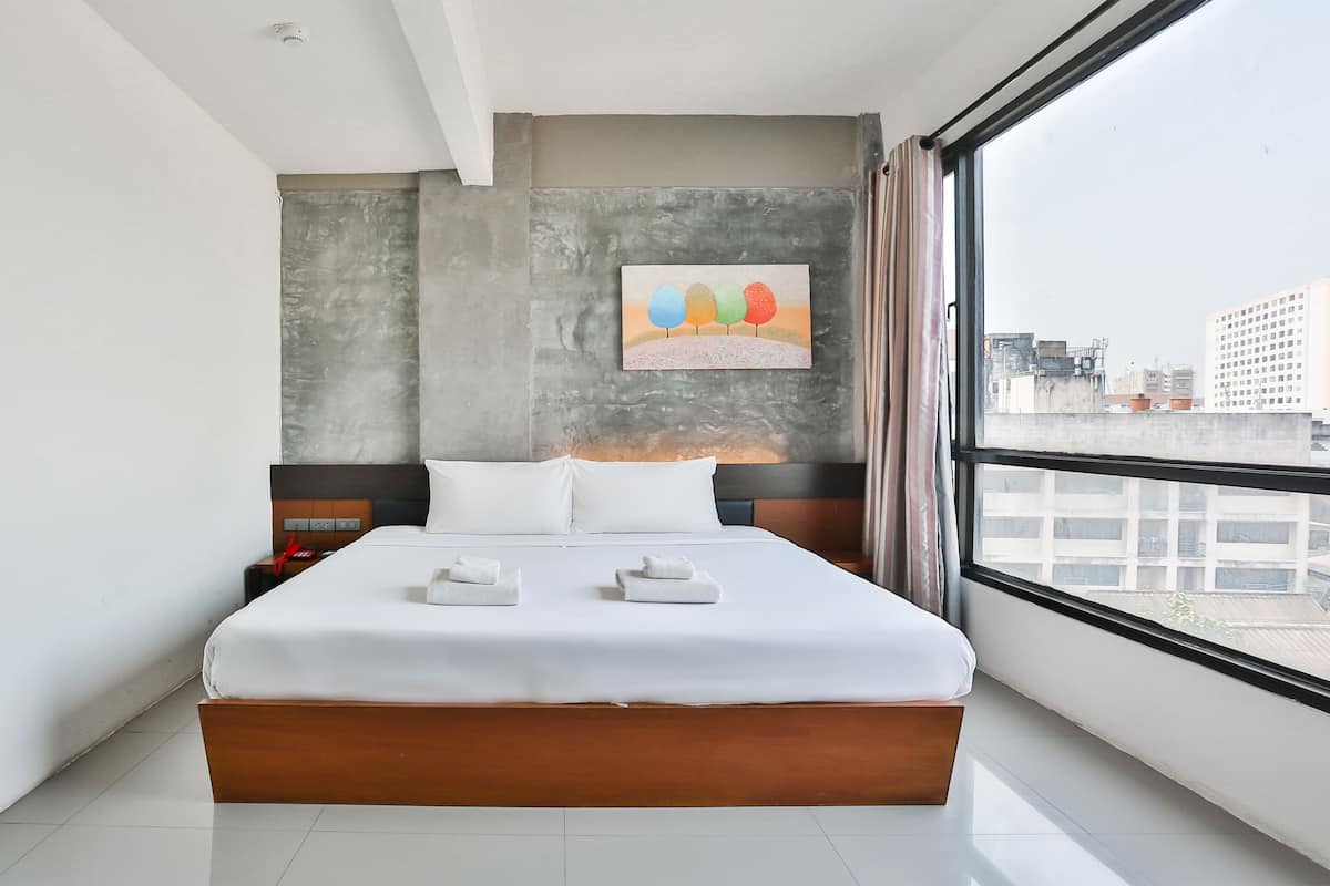 Habitació d'hotel moderna amb un llit gran, roba de llit blanca, un tabernacle de fusta, una tauleta de nit i un objecte vermell a la tauleta, amb vista a la ciutat a través d'una finestra gran.