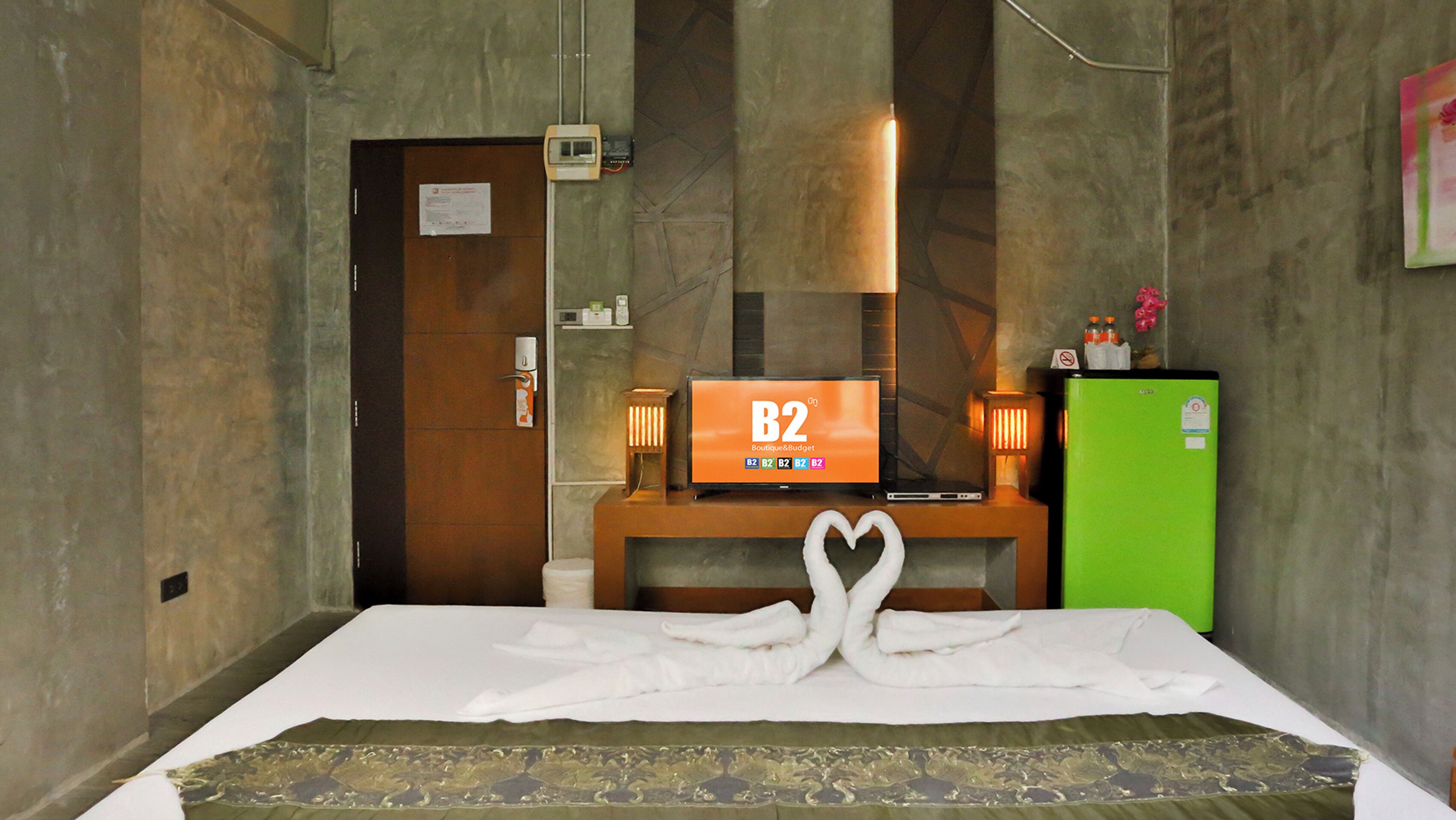 A hotel room with a bed, a television on a wooden stand, a green refrigerator, and a wall-mounted light.