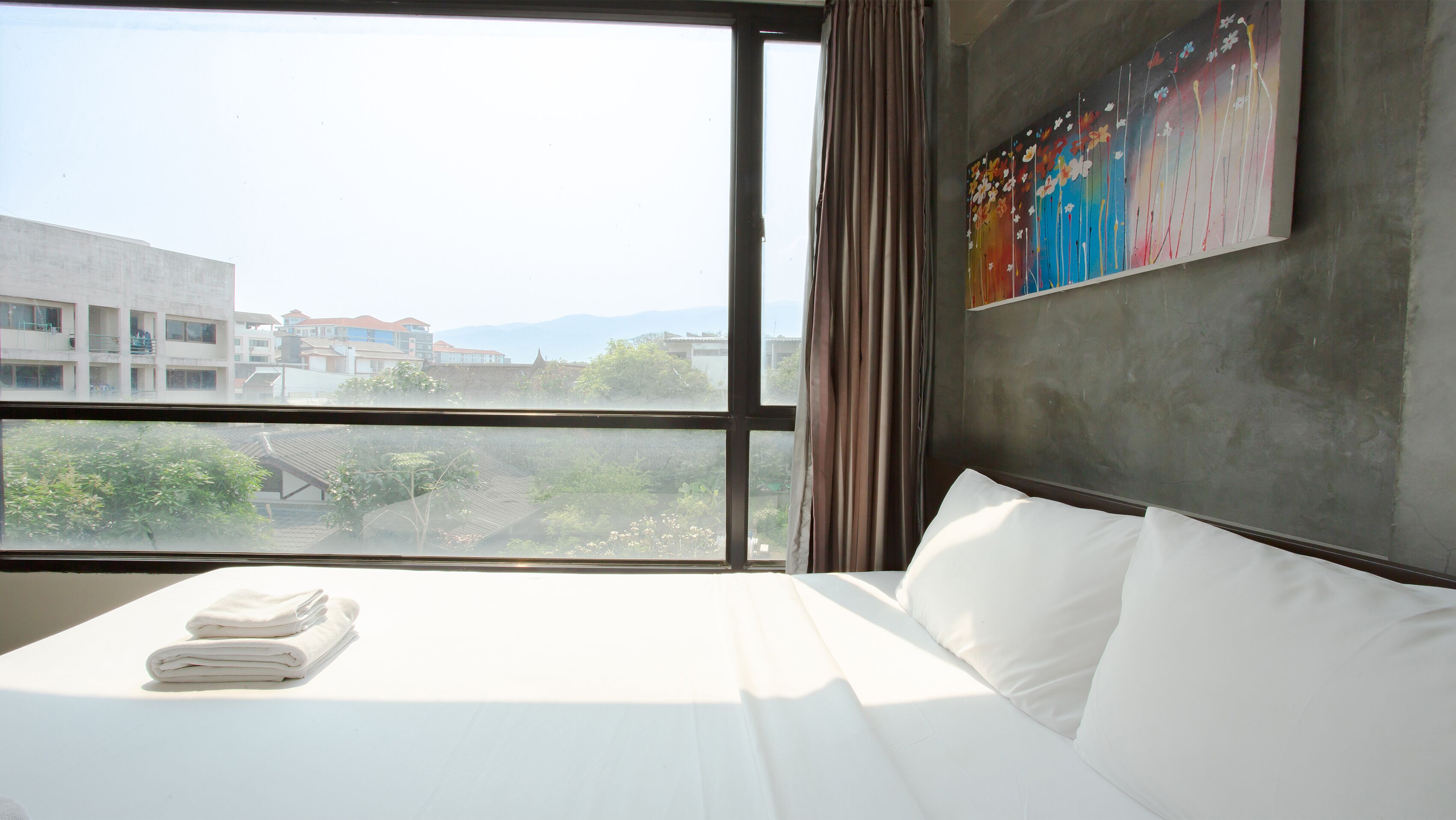 Ein Hotelzimmer mit großem Fenster, einem Bett mit weißen Leinen und einem farbenfrohen abstrakten Gemälde an der Wand.