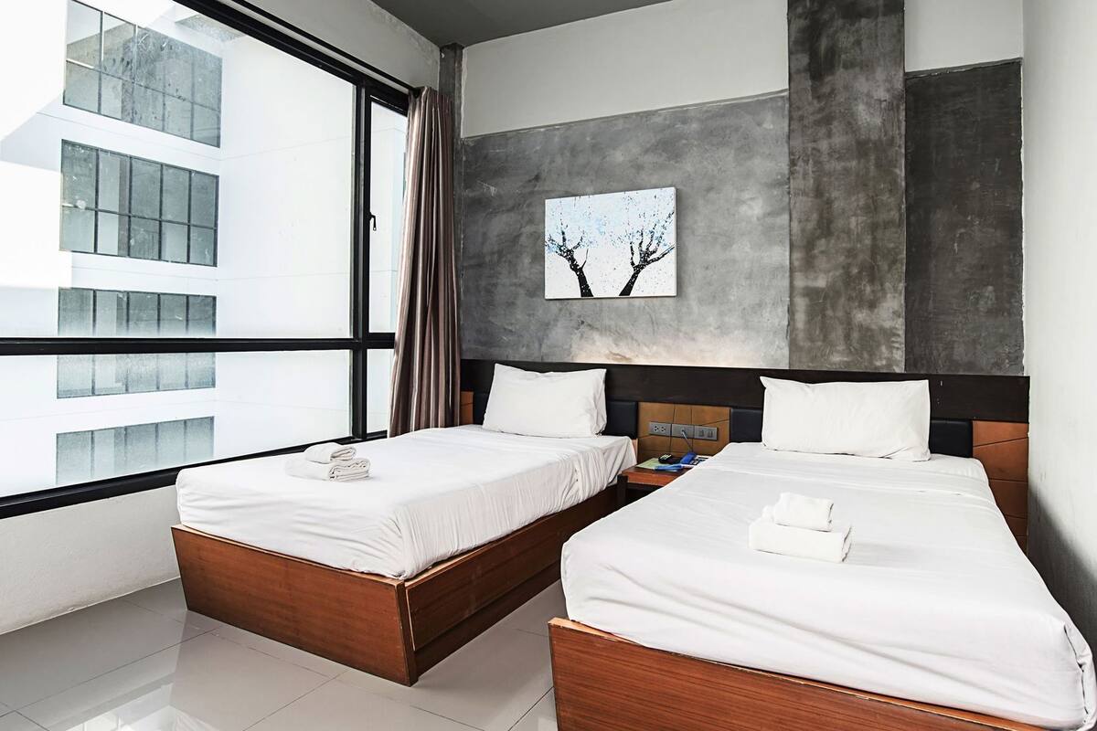 Habitació d'hotel moderna amb dos llits, una finestra gran i vista d'un edifici.