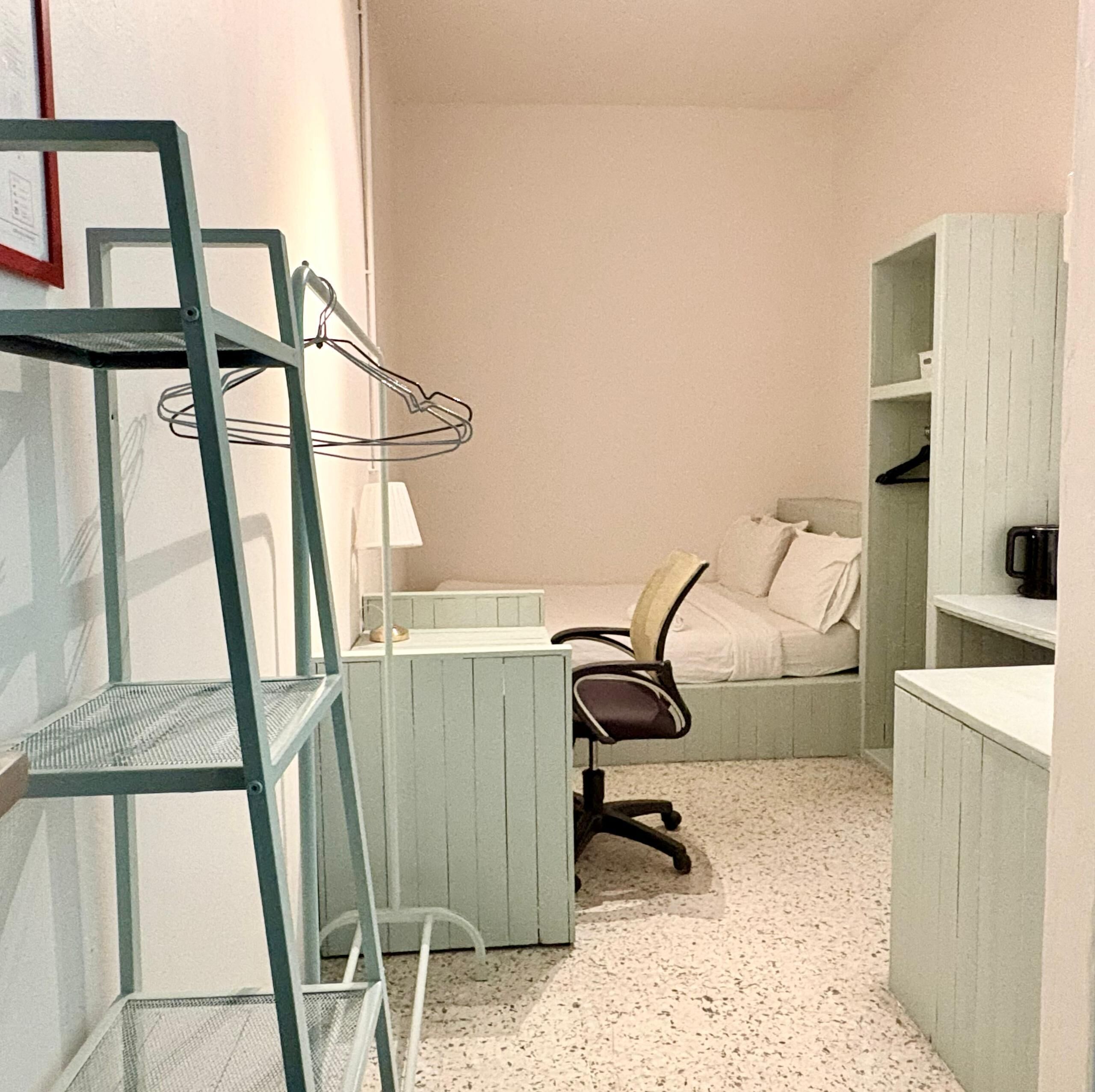 Une petite pièce, de taille modeste, comprenant un lit, un bureau et une chaise. On y trouve également des étagères et une lampe.