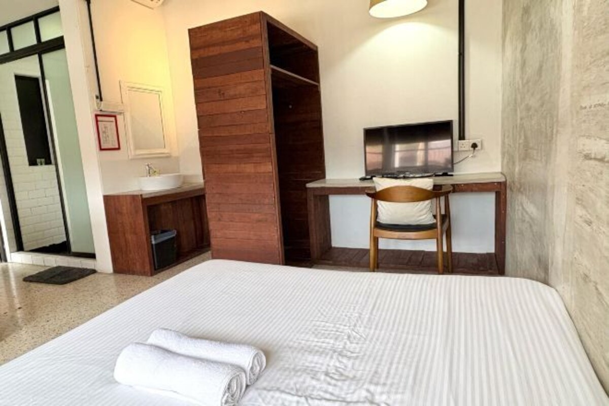 A hotel room with a large bed, a wooden wardrobe, a desk with a chair, a television, and a bathroom with a sink and mirror.