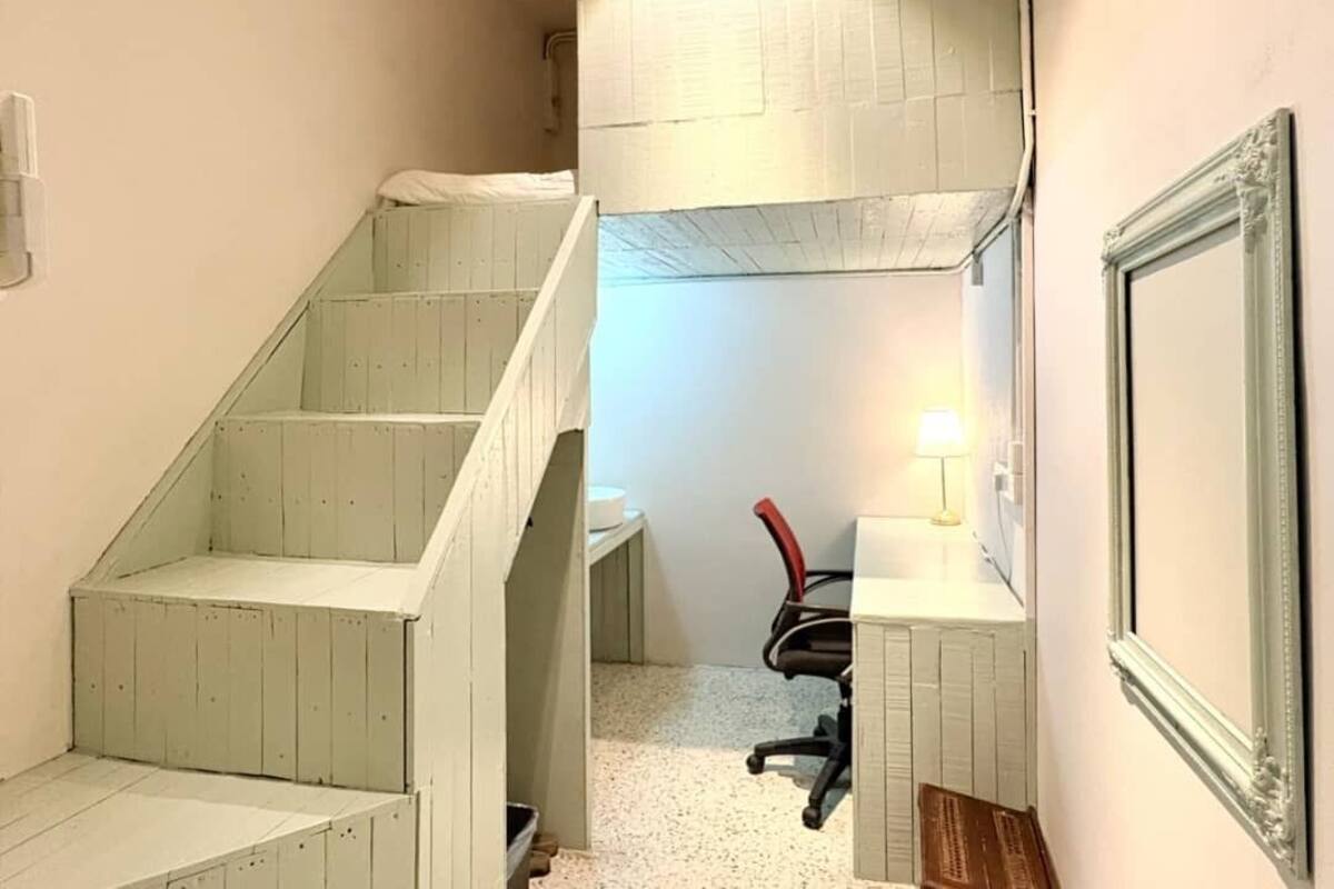 Un couloir étroit avec un escalier menant à une chambre. On peut apercevoir un bureau avec une chaise et une lampe dans la pièce.