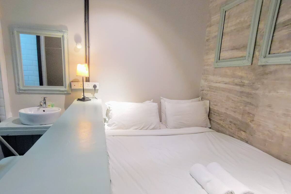 Une chambre d’hôtel avec un lit blanc, des oreillers blancs, un lavabo blanc et un miroir.