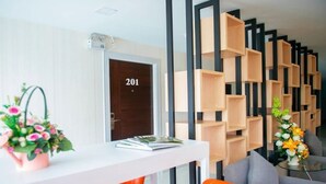 A modern hotel reception area with a white reception desk, orange chairs, and a grey sofa. There is a wooden wall with shelves and a door marked '201'. - Familyhotel (Khon Kaen)