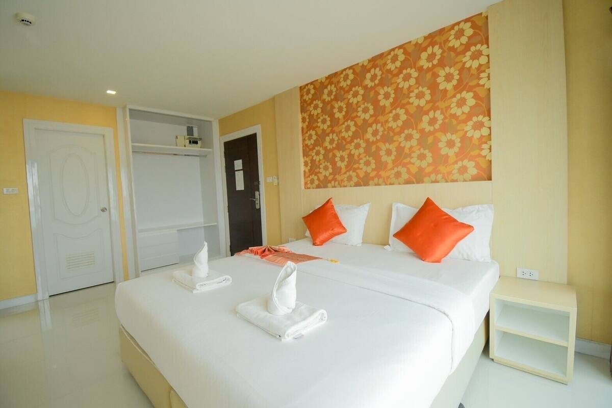A hotel room with a large bed, white bedding, orange pillows, a floral wall tapestry, and a white wardrobe.