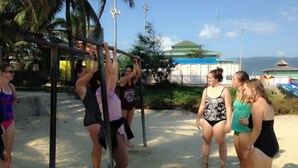 Un grupo de personas haciendo ejercicio en equipos de gimnasio al aire libre, cerca de una playa con palmeras y un edificio al fondo.