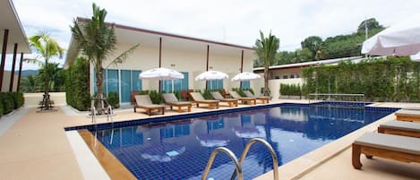 Une piscine entourée de verdure, agrémentée de chaises longues et de parasols, et bordée d’un bâtiment dont le toit est recouvert de tuiles.